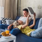 young couple sitting on sofa at home looking in tablet, watching online, using internet, smiling, happy together, having fun, man and woman in love, family leisure time, relaxing, modern devices